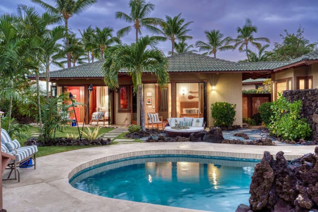 An evening view of a luxury tropical villa with a private swimming pool, palm trees, and illuminated open bedrooms.