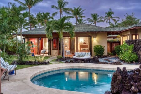 An evening view of a luxury tropical villa with a private swimming pool, palm trees, and illuminated open bedrooms.