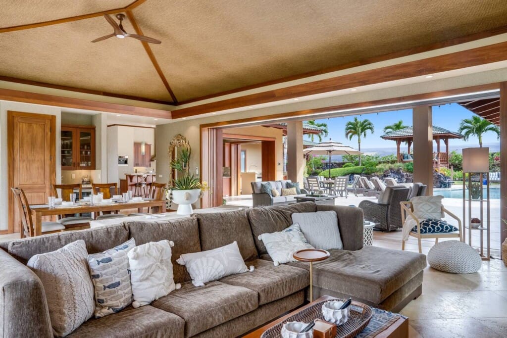 An open-concept living room with a large brown sectional sofa, a vaulted woven ceiling, and sliding glass pocket doors that open completely to an outdoor patio pool area.