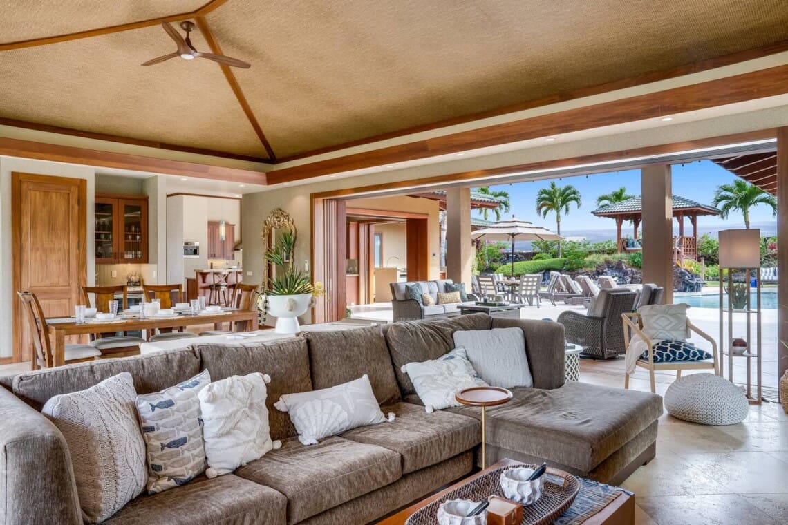 An open-concept living room with a large brown sectional sofa, a vaulted woven ceiling, and sliding glass pocket doors that open completely to an outdoor patio pool area.