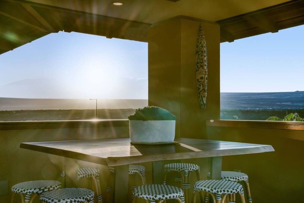A sun-drenched view of an upstairs covered lanai featuring a tall wooden bar table with woven barstools and an open-air railing showcasing a bright horizon.