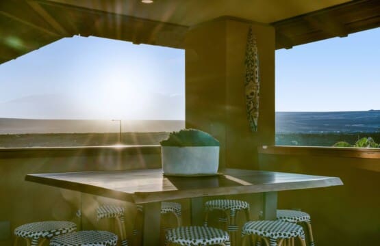 A sun-drenched view of an upstairs covered lanai featuring a tall wooden bar table with woven barstools and an open-air railing showcasing a bright horizon.