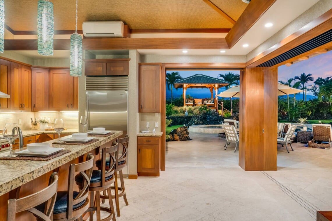 A modern kitchen with wooden cabinetry, a granite breakfast bar, and large pocket doors opening completely to an outdoor patio with a pavilion at dusk.