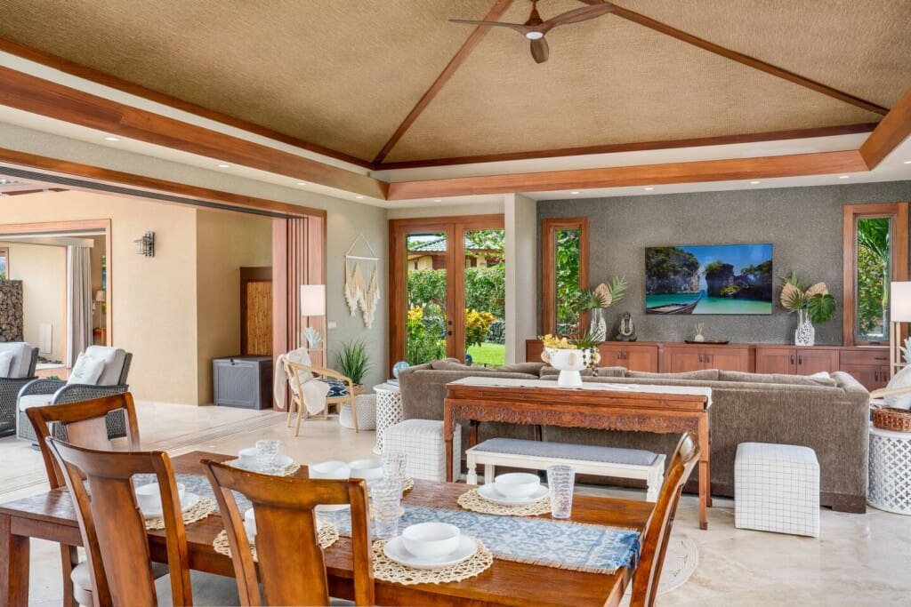 An open-air living room and dining area with high woven ceilings, a wooden dining table, a large sectional sofa, and a wall-mounted TV.