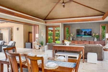An open-air living room and dining area with high woven ceilings, a wooden dining table, a large sectional sofa, and a wall-mounted TV.