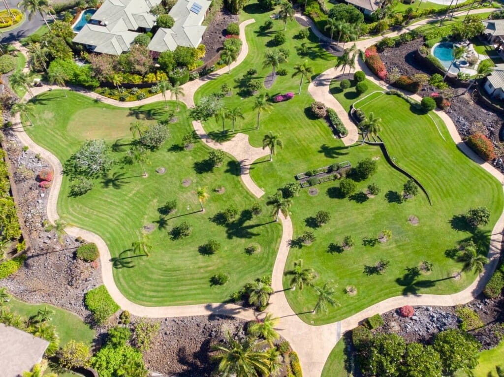 An aerial view of a lush community park featuring green lawns, winding paved walking paths, palm trees, and organized garden beds.