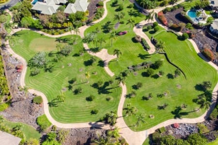 An aerial view of a lush community park featuring green lawns, winding paved walking paths, palm trees, and organized garden beds.