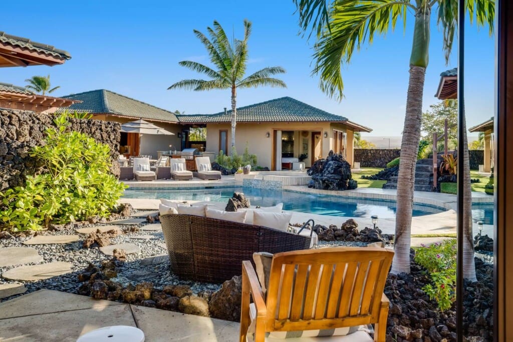 A view looking out from a doorway toward a luxury backyard courtyard featuring a custom swimming pool, outdoor lounge furniture, volcanic rock landscaping, and pod-style villa structures under a bright blue sky.