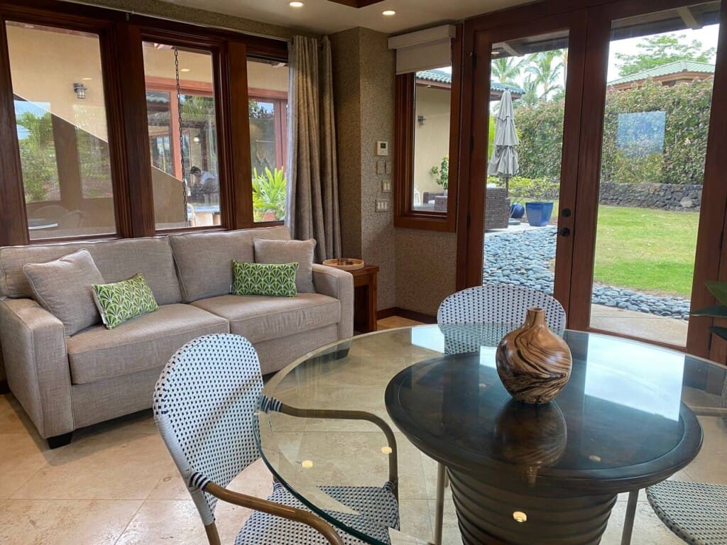 A bright game room featuring a light grey sofa with green patterned pillows, a round glass-top table with woven chairs, and large windows and doors looking out to a grassy yard.