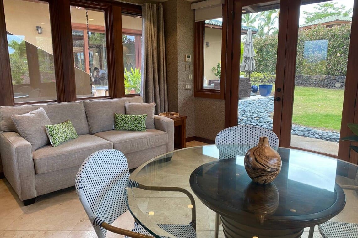 A bright game room featuring a light grey sofa with green patterned pillows, a round glass-top table with woven chairs, and large windows and doors looking out to a grassy yard.