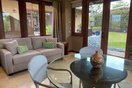 A bright game room featuring a light grey sofa with green patterned pillows, a round glass-top table with woven chairs, and large windows and doors looking out to a grassy yard.