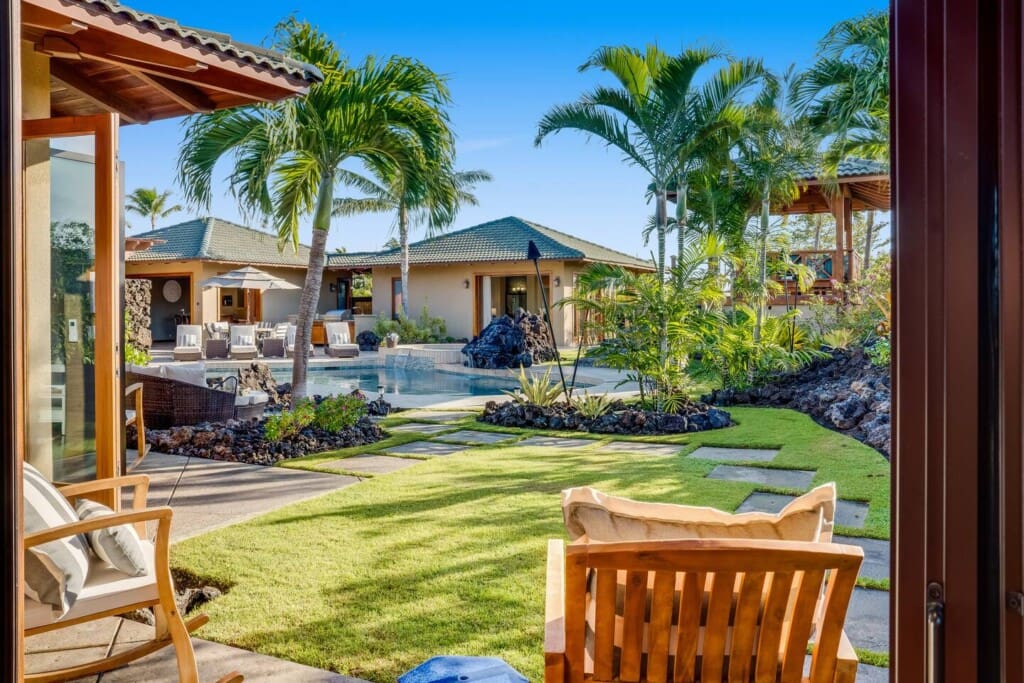 Alt Text An outdoor perspective looking out from an open doorway onto a manicured green lawn with a stone walking path, palm trees, and a swimming pool in the background.