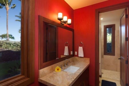 A modern bathroom vanity with a stone countertop and large wood-framed mirror set against vibrant red walls, with a window showing a palm tree outside.