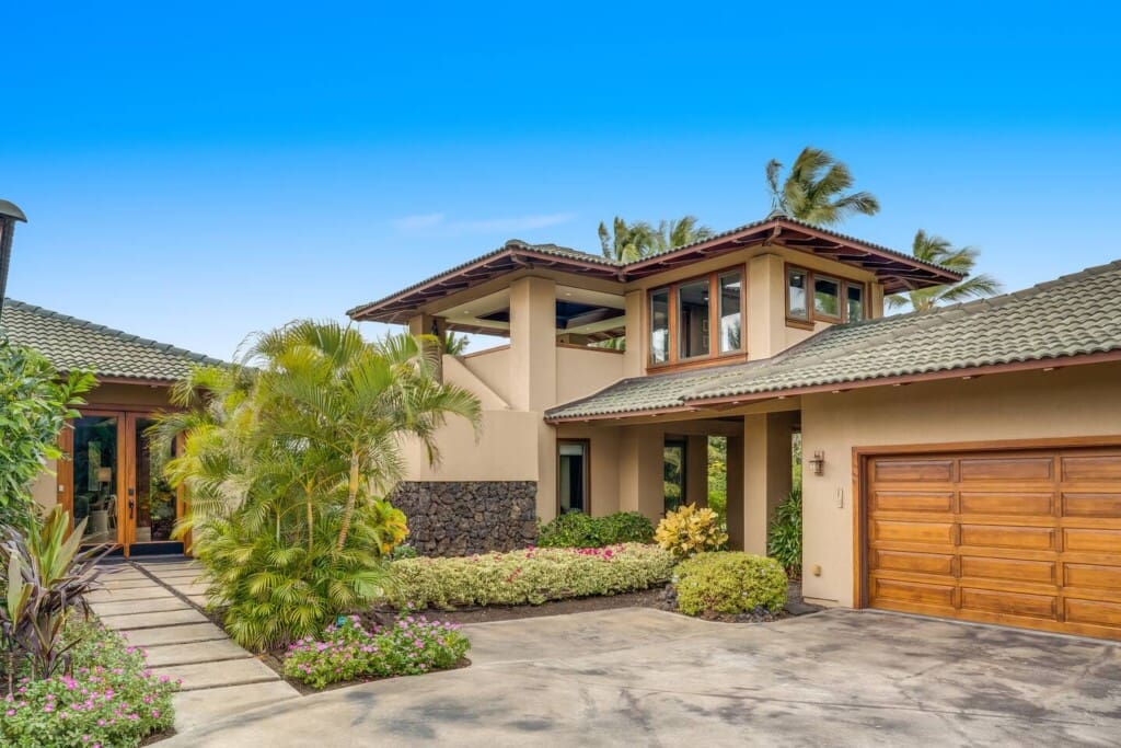 An exterior view of a luxury home showing a concrete driveway, a wooden garage door, an upstairs open-air pavilion, and a stone path leading to the front door entry.