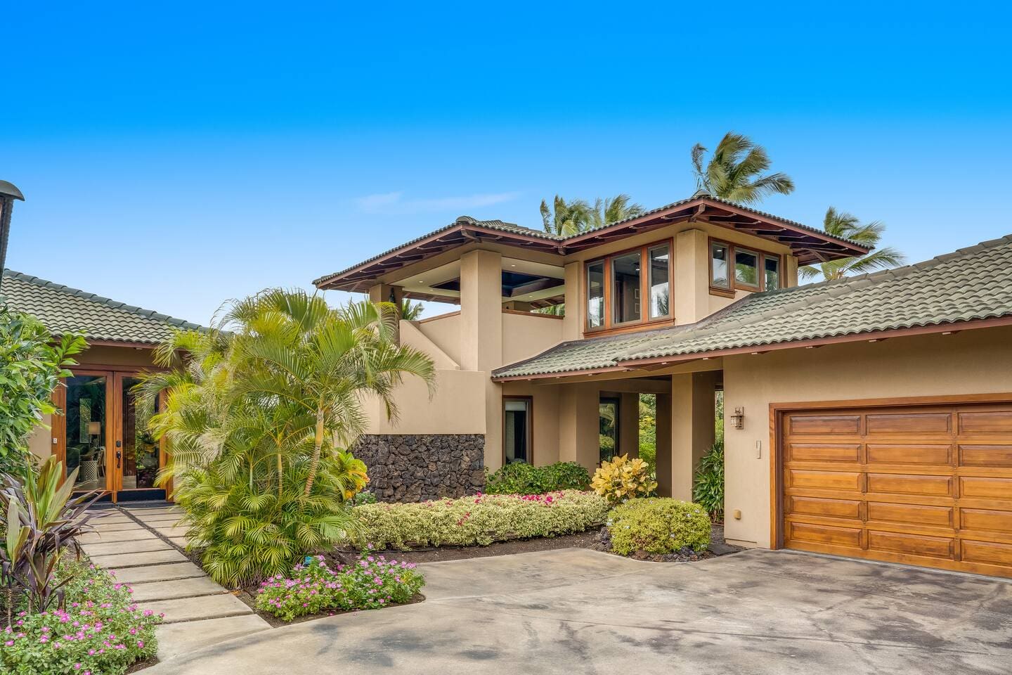 An exterior view of a luxury home showing a concrete driveway, a wooden garage door, an upstairs open-air pavilion, and a stone path leading to the front door entry.