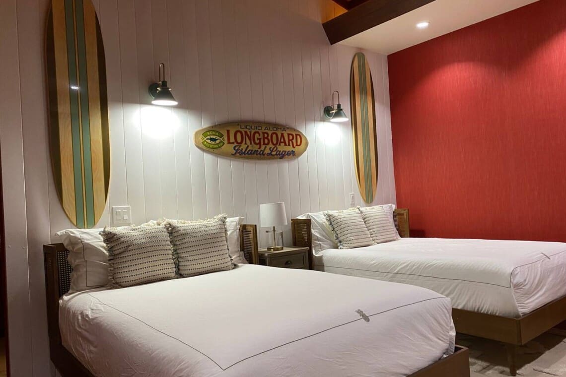 A bright bedroom featuring two beds with white linens, a red accent wall, white wood paneling, and decorative surfboards mounted above the headboards.