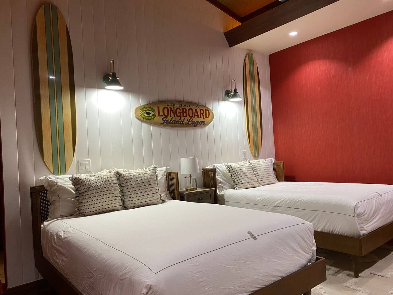 A bright bedroom featuring two beds with white linens, a red accent wall, white wood paneling, and decorative surfboards mounted above the headboards.