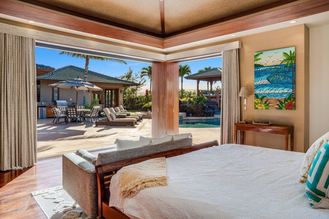 A bright bedroom with large corner pocket doors fully opened to a sunny backyard patio featuring a pool, sun loungers, and an outdoor dining area.