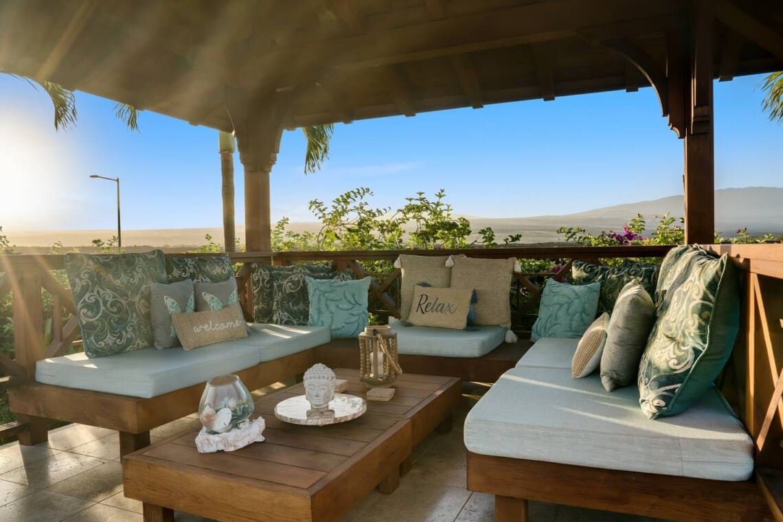 An interior view of a covered wooden gazebo featuring comfortable wrap-around cushioned bench seating, decorative pillows, and a low wooden coffee table under a clear blue sky.