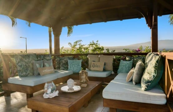 An interior view of a covered wooden gazebo featuring comfortable wrap-around cushioned bench seating, decorative pillows, and a low wooden coffee table under a clear blue sky.