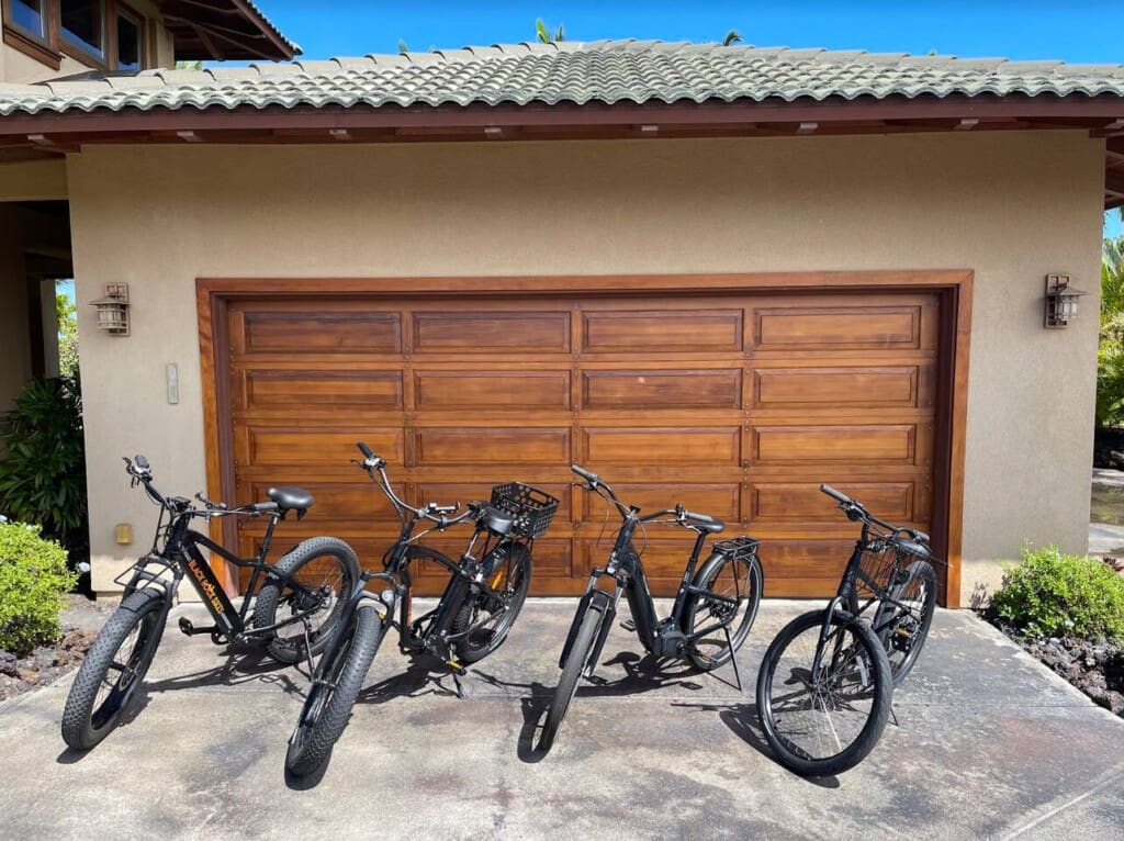Four black bicycles, including electric bikes and a standard bicycle, parked side-by-side on a concrete driveway in front of a wooden garage door.