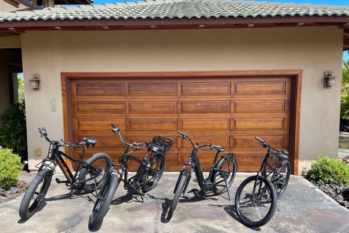 Four black bicycles, including electric bikes and a standard bicycle, parked side-by-side on a concrete driveway in front of a wooden garage door.