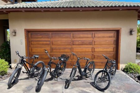 Four black bicycles, including electric bikes and a standard bicycle, parked side-by-side on a concrete driveway in front of a wooden garage door.