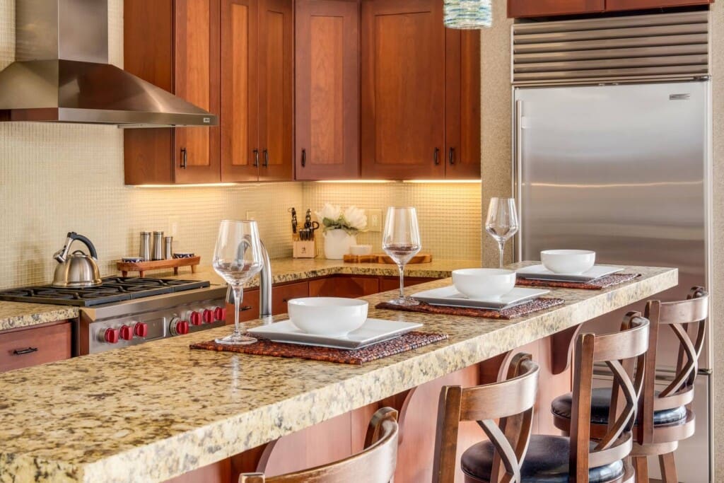 A close-up view of a gourmet kitchen breakfast bar set with plates and wine glasses, featuring wood cabinetry, granite countertops, and modern stainless steel appliances.