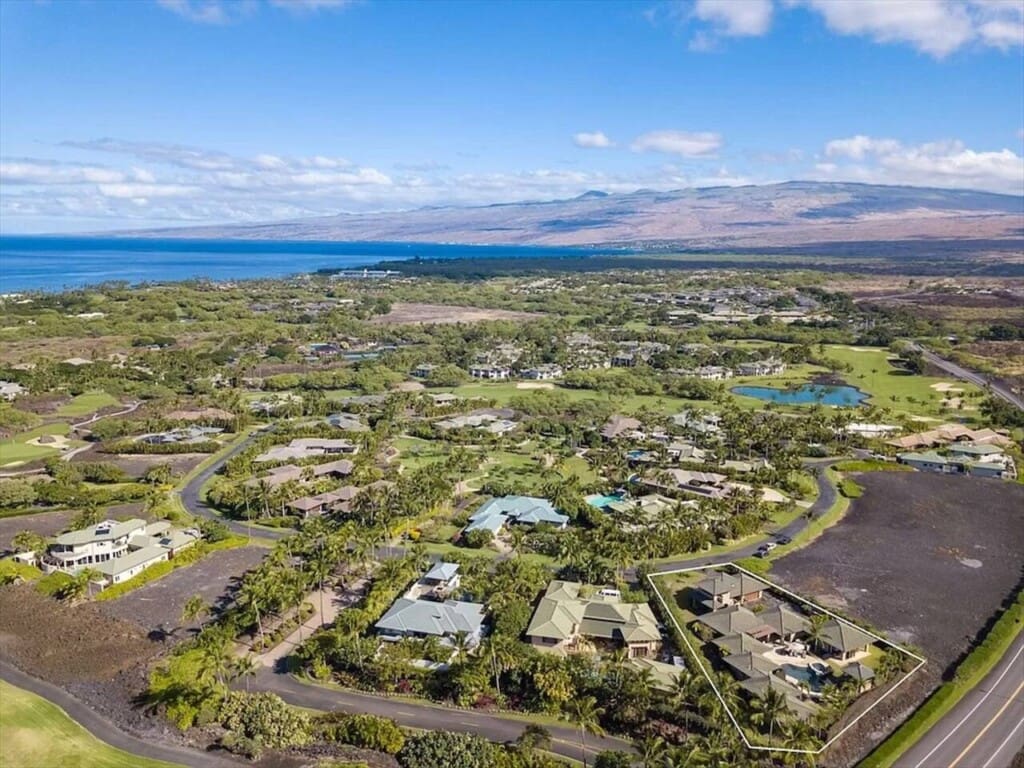 An aerial view of a luxury residential community showing a highlighted estate surrounded by tropical landscaping, roads, dark volcanic rock terrain, and an ocean backdrop under a blue sky.