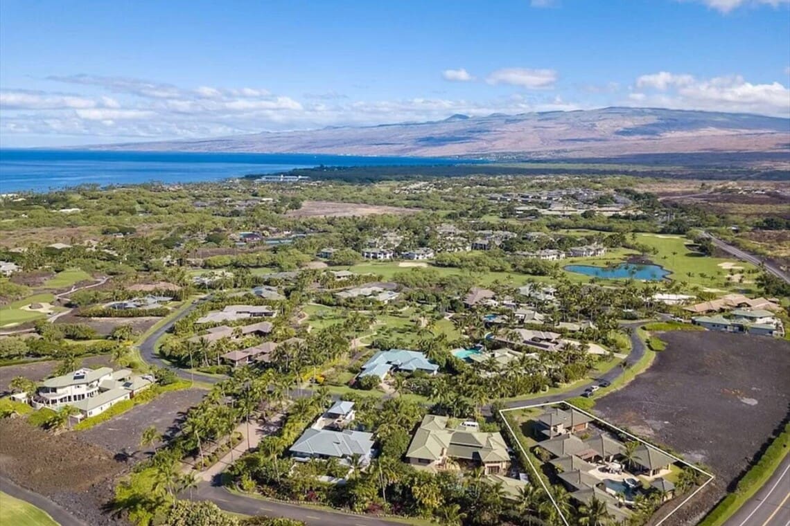 An aerial view of a luxury residential community showing a highlighted estate surrounded by tropical landscaping, roads, dark volcanic rock terrain, and an ocean backdrop under a blue sky.