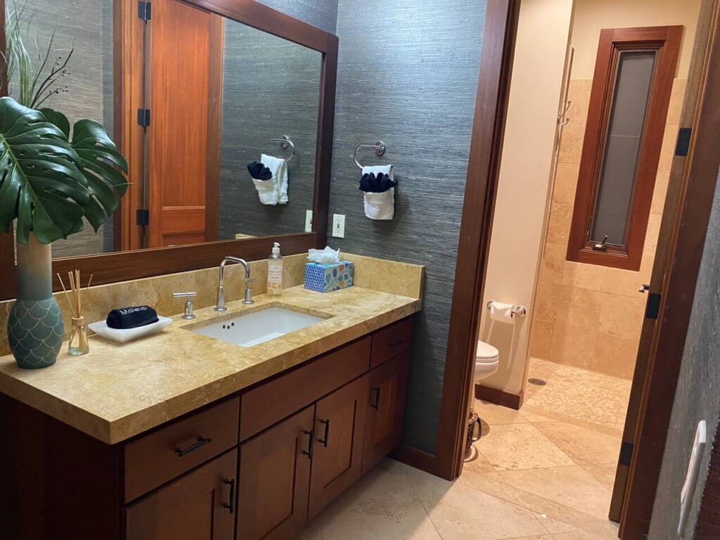 A modern bathroom featuring a wooden vanity with a stone countertop, a large mirror, dark textured wallpaper, and an adjacent walk-in shower area.