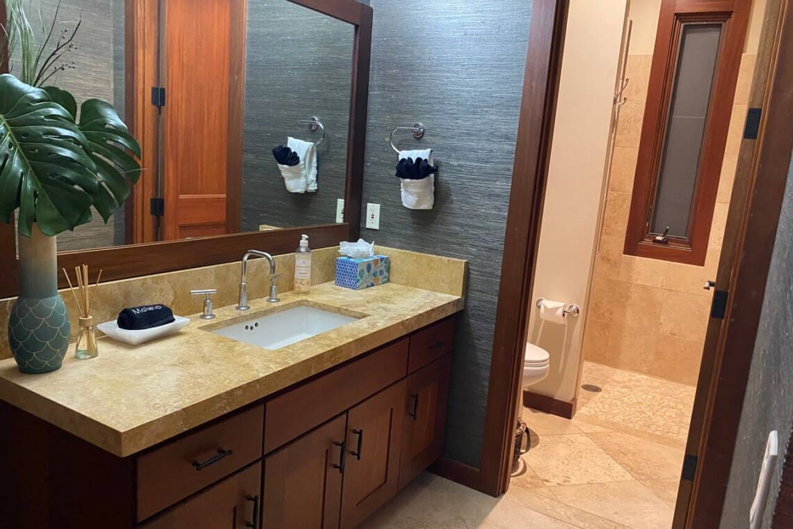 A modern bathroom featuring a wooden vanity with a stone countertop, a large mirror, dark textured wallpaper, and an adjacent walk-in shower area.