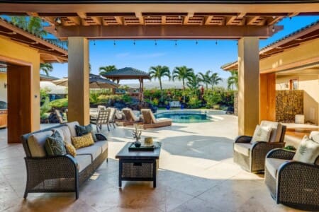 An outdoor covered patio with a wicker sofa and armchairs looking out onto a private swimming pool, sun loungers, and a backyard gazebo.