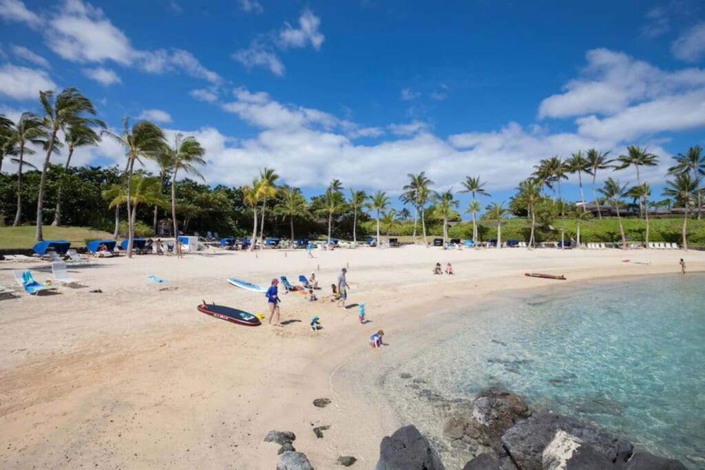 A sunny white sand beach with clear turquoise water, fringed by palm trees, blue beach cabanas, and people relaxing near the shore under a bright blue sky.