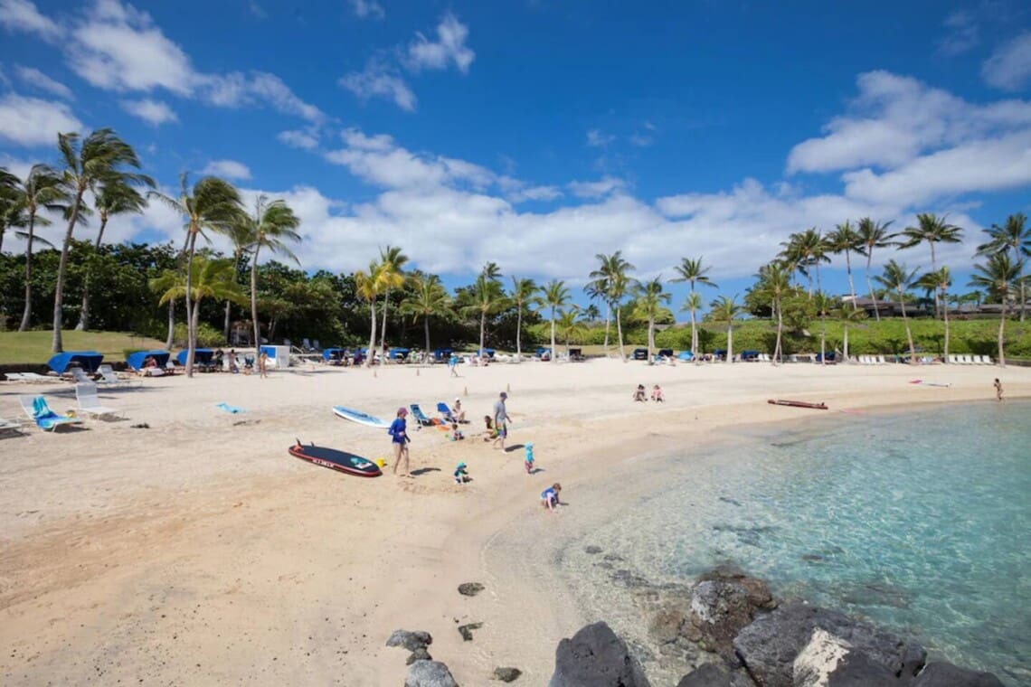 A sunny white sand beach with clear turquoise water, fringed by palm trees, blue beach cabanas, and people relaxing near the shore under a bright blue sky.