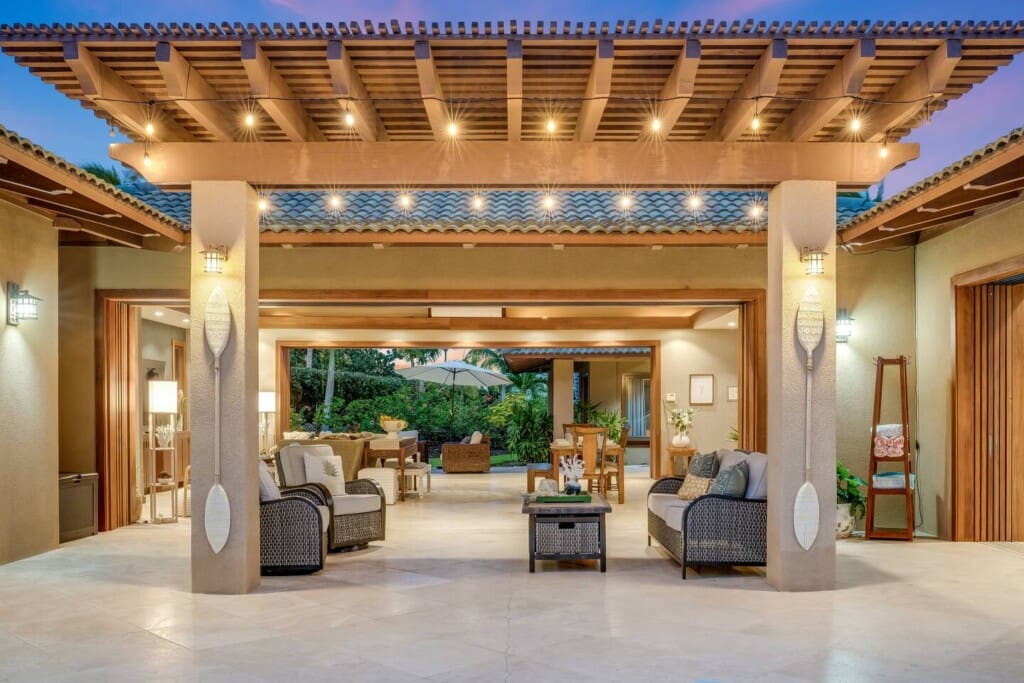 An outdoor covered sitting area with string lights looking through fully opened pocket doors into a living room and dining area at twilight.