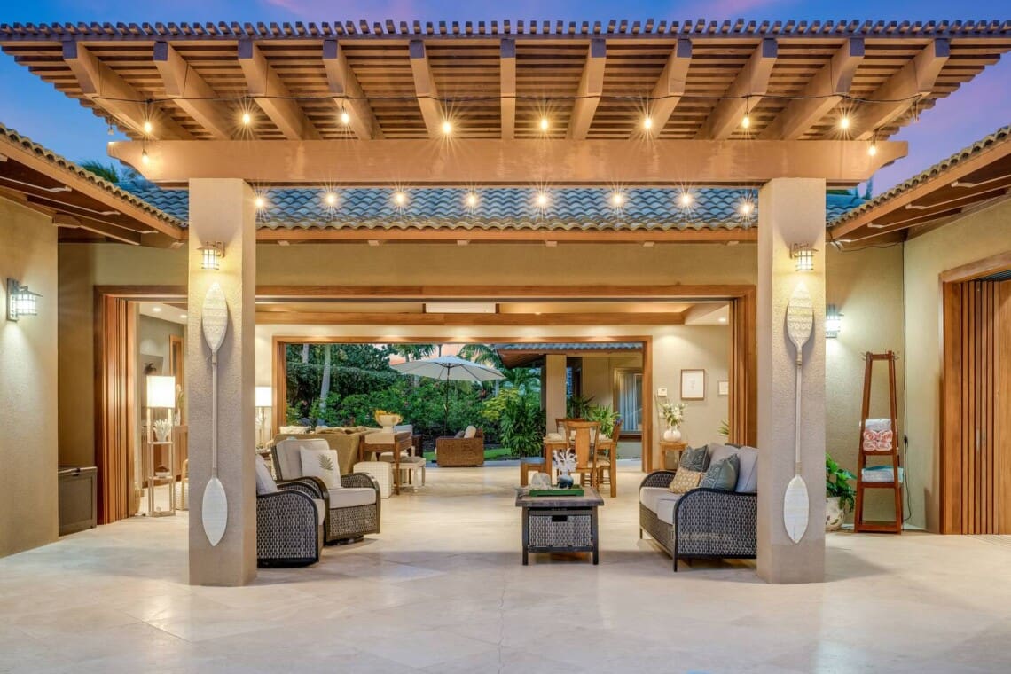 An outdoor covered sitting area with string lights looking through fully opened pocket doors into a living room and dining area at twilight.