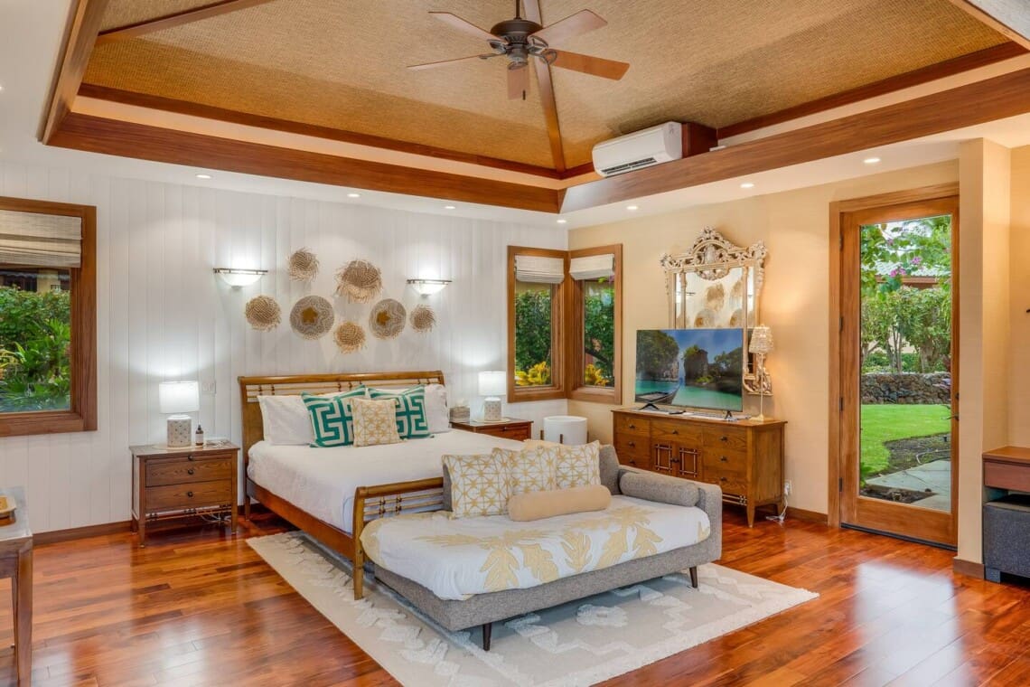 A spacious tropical bedroom with a wooden bed frame, a daybed at the foot of the bed, hardwood floors, and a woven ceiling with a ceiling fan.