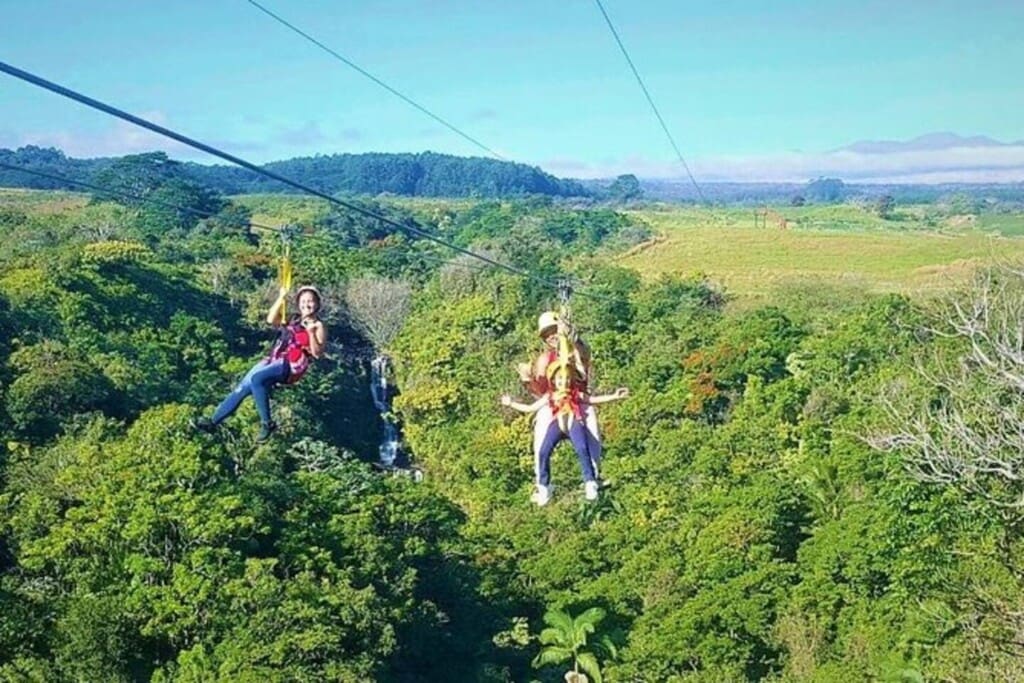 People ziplining side-by-side high above a lush, green tropical forest canopy with a waterfall visible in the background under a blue sky.