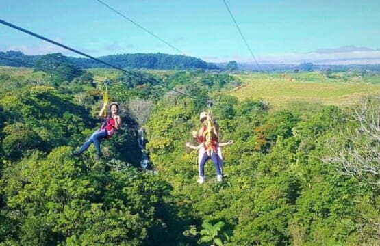 People ziplining side-by-side high above a lush, green tropical forest canopy with a waterfall visible in the background under a blue sky.