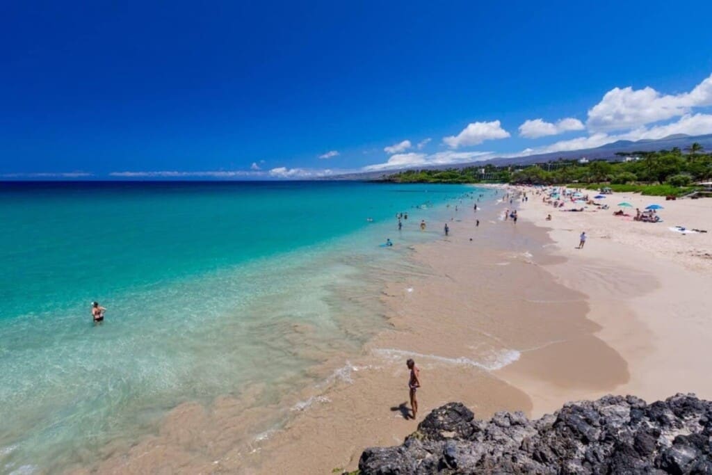 An elevated view looking along a wide sandy beach next to crystal-clear turquoise ocean water with swimmers under a bright blue sky.