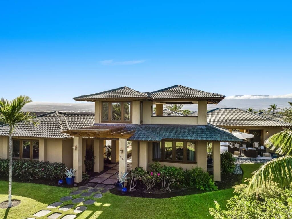 A luxury multi-level estate with tan stucco walls, a dark tiled roof, a manicured green lawn with a stone stepping path, and a scenic mountain backdrop under a clear blue sky.