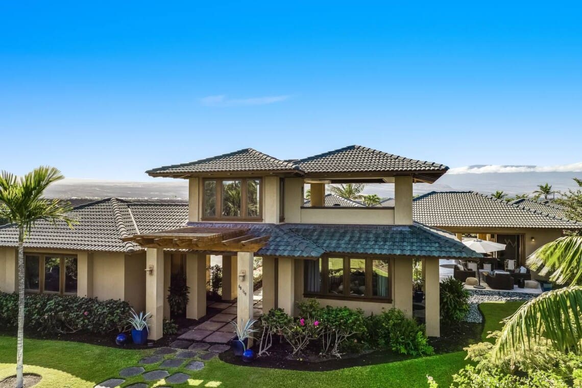 A luxury multi-level estate with tan stucco walls, a dark tiled roof, a manicured green lawn with a stone stepping path, and a scenic mountain backdrop under a clear blue sky.