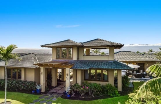 A luxury multi-level estate with tan stucco walls, a dark tiled roof, a manicured green lawn with a stone stepping path, and a scenic mountain backdrop under a clear blue sky.