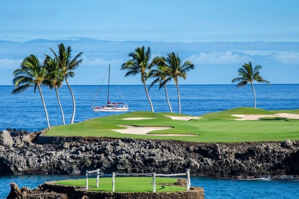 A scenic view of a green golf course green on a rocky lava shoreline with palm trees, sand bunkers, and a sailboat on the blue ocean in the background.