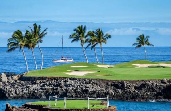 A scenic view of a green golf course green on a rocky lava shoreline with palm trees, sand bunkers, and a sailboat on the blue ocean in the background.