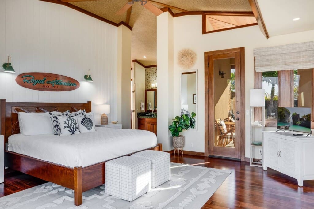 A spacious primary bedroom with a wooden bed frame, white bedding with coral-patterned pillows, a vaulted woven ceiling, and a wooden glass door leading outside.
