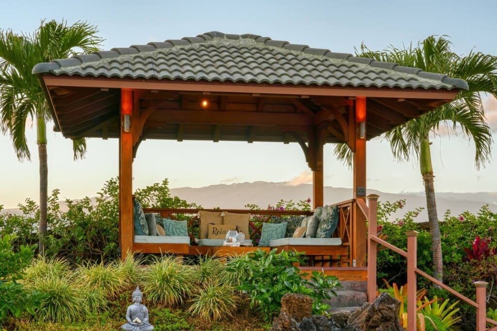 An elevated outdoor wooden gazebo with cushioned seating surrounded by tropical plants and palm trees, overlooking distant mountain views.