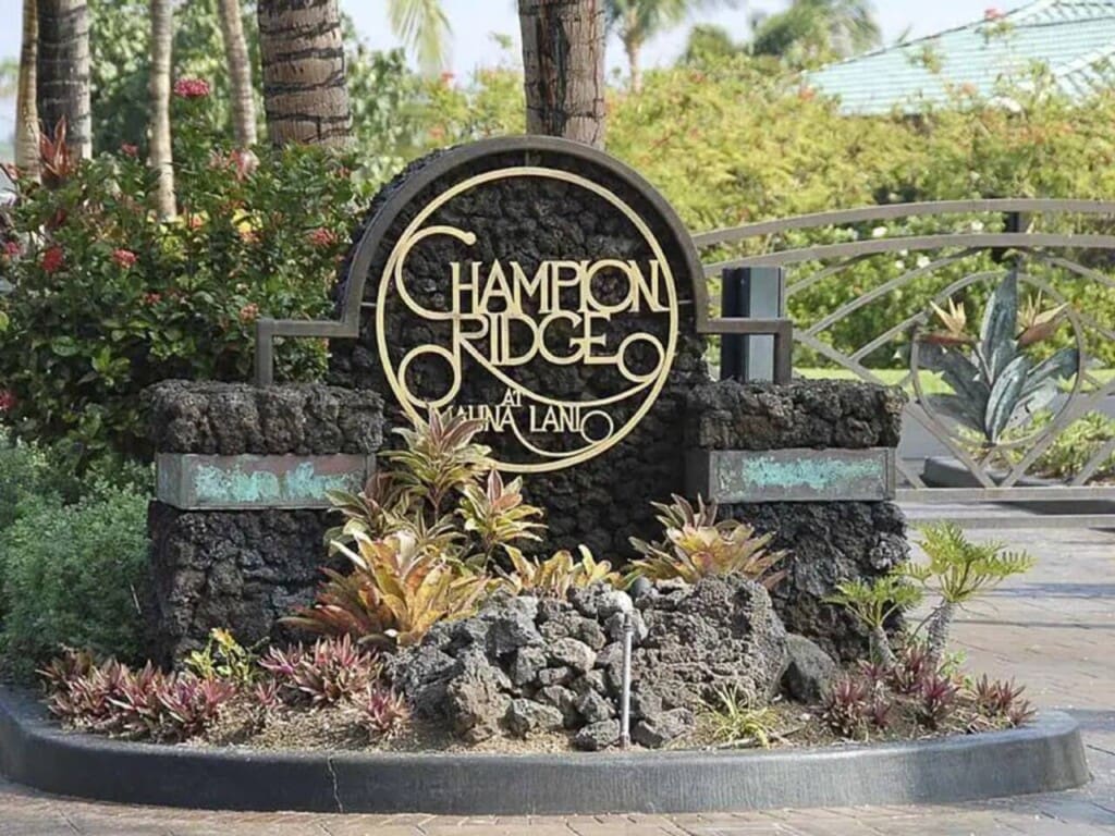 A large stone monument sign reading "Champion Ridge at Mauna Lani" surrounded by volcanic rocks and tropical landscaping at a gated entrance.