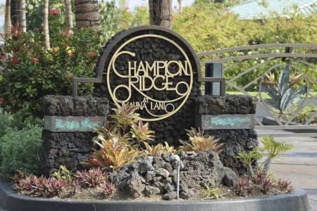 A large stone monument sign reading "Champion Ridge at Mauna Lani" surrounded by volcanic rocks and tropical landscaping at a gated entrance.